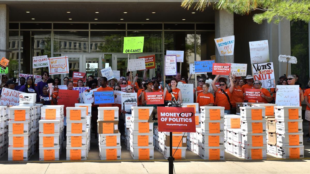 A crowd of supporters with signs stands behind rows of filing boxes on a curb