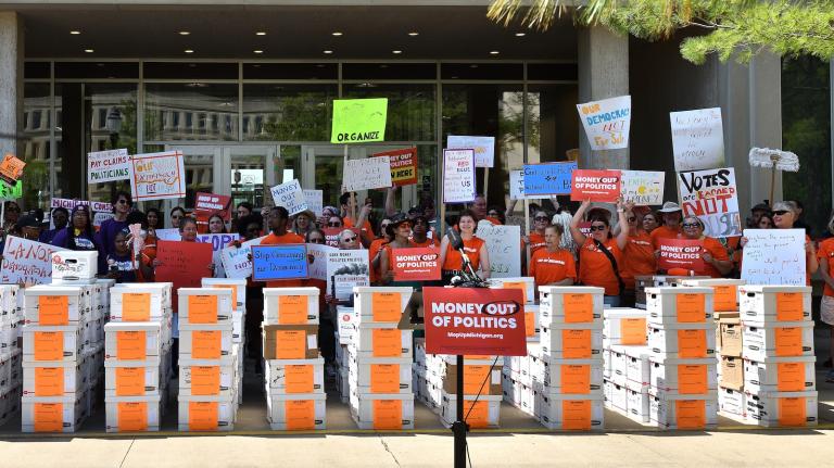 A crowd of supporters with signs stands behind rows of filing boxes on a curb