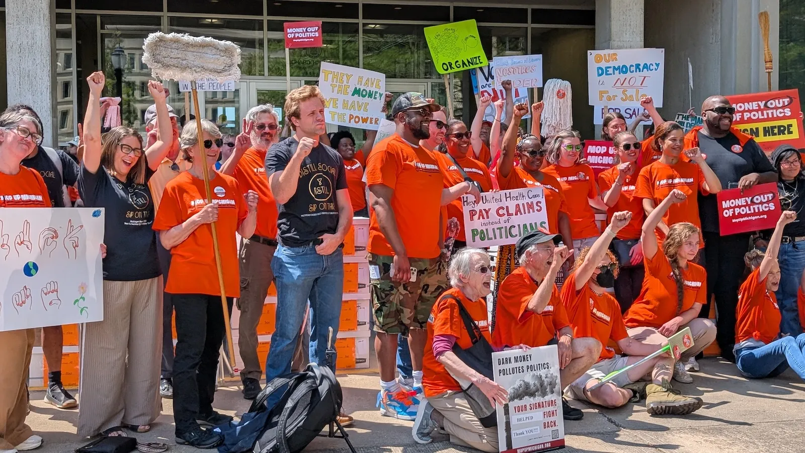Supporters in matching orange tee shirts carry signs on a sidewalk
